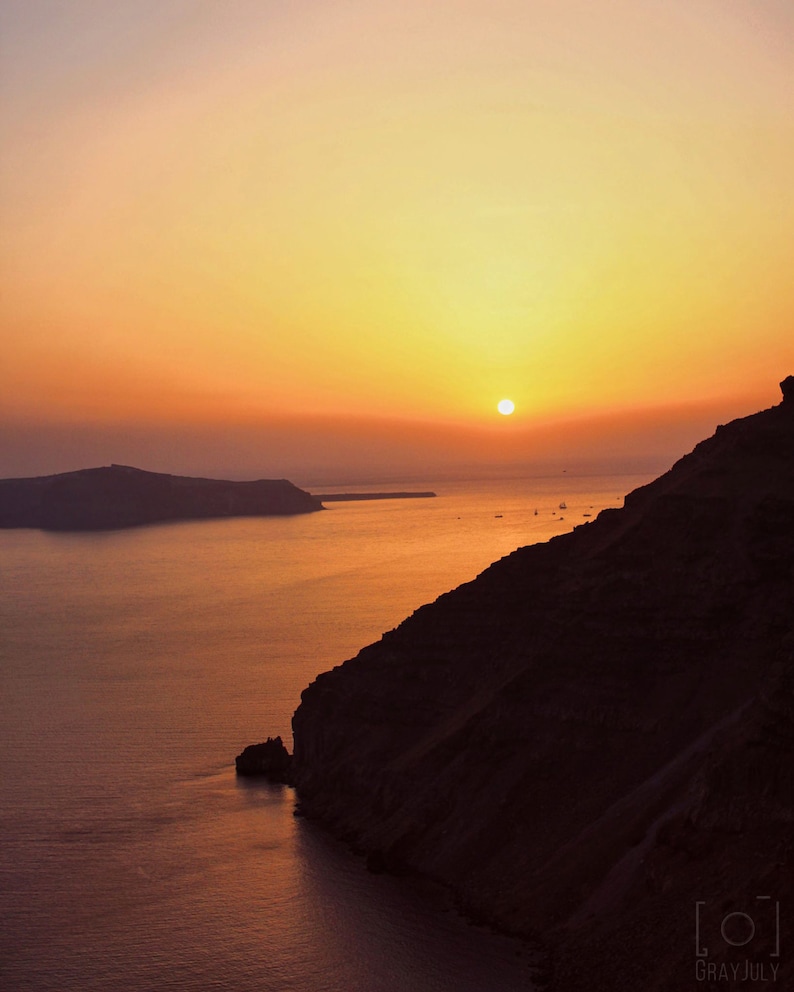 May include: A scenic sunset over a body of water with a large, dark cliff in the foreground. The sun is setting behind a distant island, casting a warm orange glow over the scene.