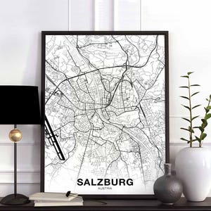 May include: A black and white map of Salzburg, Austria, framed in black. The map details the city's streets and landmarks. The word "SALZBURG" and "AUSTRIA" are printed at the bottom. A black lamp and white vase are on the table.