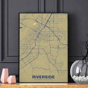 May include: Framed map print of Riverside, California, featuring a detailed street and road network in navy blue against a tan background. The city name and state are printed in navy blue at the bottom. The print is displayed on a wooden shelf with decorative vases.