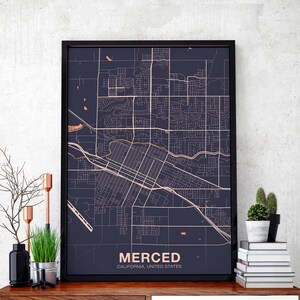 May include: A black framed print of a map of Merced, California, United States. The map is in a dark blue color with white lines showing the streets and roads. The city name is printed in a light orange color at the bottom of the map.