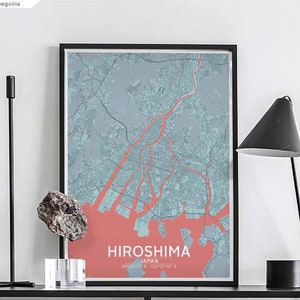 Puede incluir: Un mapa enmarcado de la ciudad de Hiroshima, Japón, con un esquema de color azul claro y rojo. El mapa muestra las calles y vías fluviales de la ciudad. El texto "HIROSHIMA JAPÓN 34°23'07" N 132°27'19" E" está impreso en la parte inferior del mapa.