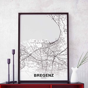 May include: A black and white map print of Bregenz, Austria, in a black frame. The map shows the city's streets and waterways. The print is hanging on a white wall with two black candle holders and a white vase with green plants.