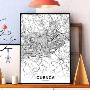 May include: Black and white line art map print of Cuenca, Ecuador. The print is framed and hanging on a wall with a wooden shelf below it.