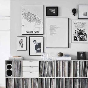 Puede incluir: Una estanter&iacute;a blanca con varios estantes llenos de discos de vinilo. La estanter&iacute;a est&aacute; contra una pared blanca con obras de arte enmarcadas en negro, incluyendo un mapa de Puerto Plata, una foto en blanco y negro, un poema y un p&oacute;ster con el texto "Rocket Crypt".