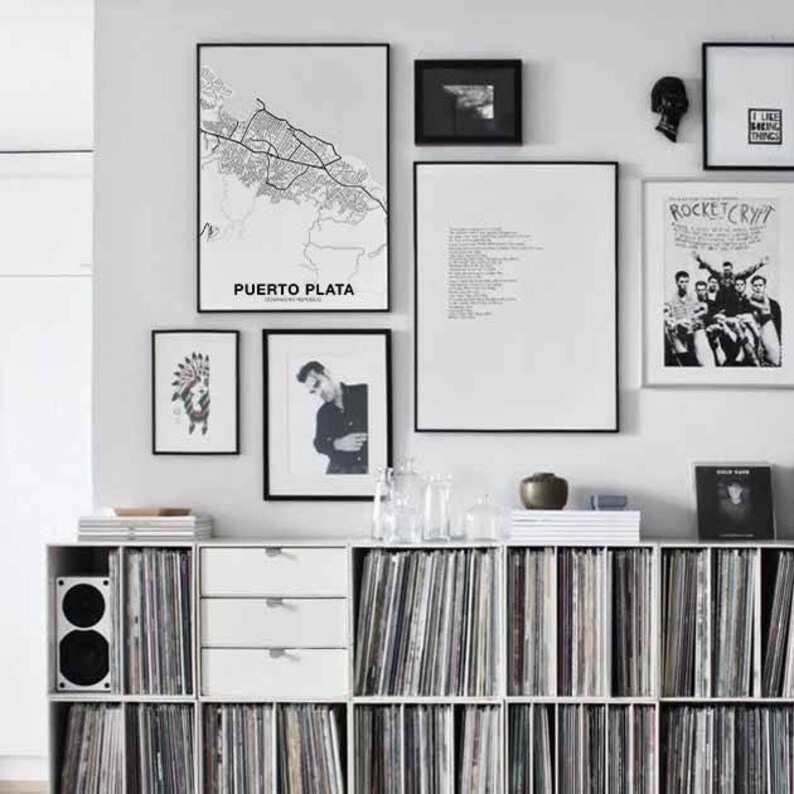 Puede incluir: Una estanter&iacute;a blanca con varios estantes llenos de discos de vinilo. La estanter&iacute;a est&aacute; contra una pared blanca con obras de arte enmarcadas en negro, incluyendo un mapa de Puerto Plata, una foto en blanco y negro, un poema y un p&oacute;ster con el texto "Rocket Crypt".