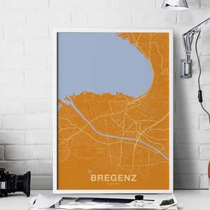May include: A map print of Bregenz, Austria, with a light blue lake and orange landmass. The city streets are shown in white lines. The print is framed in a white frame.