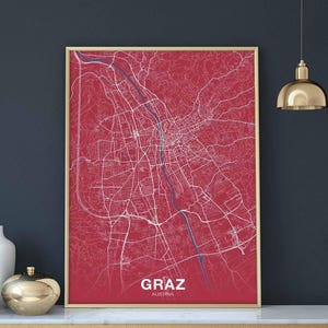 May include: A red and white map of Graz, Austria, with the city name and country printed at the bottom. The map is framed in a gold frame and is hanging on a dark blue wall.