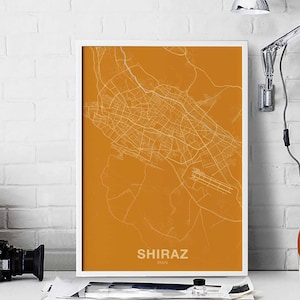 Op de afbeelding: Een witte ingelijste print van een kaart van Shiraz, Iran. De kaart is in witte lijnen op een effen oranje achtergrond. De stadsnaam "SHIRAZ" en "IRAN" zijn in wit onderaan de kaart gedrukt.