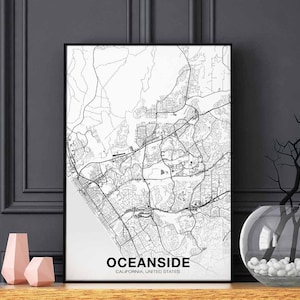 Puede incluir: Una impresión de mapa en blanco y negro de Oceanside, California, Estados Unidos. El mapa muestra las calles y los puntos de referencia de la ciudad. La impresión está enmarcada en un marco negro y está colgada en una pared gris.