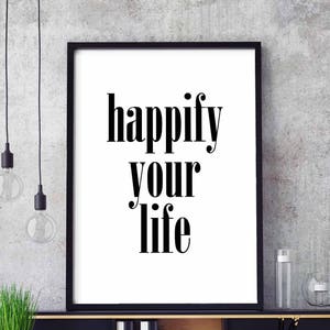 Może przedstawiać: Czarno-biały wydruk typograficzny z tekstem "happify your life" na białym tle. Wydruk jest oprawiony w czarną ramę i wisi na szarej ścianie.