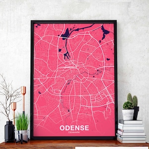 May include: A black framed print of a pink map of Odense, Denmark. The map shows the city's streets and waterways. The city name is printed in white at the bottom of the map.