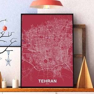 Op de afbeelding: Een rode en witte stadsplattegrondprint met het woord "TEHRAN" en "IRAN" in witte tekst onderaan. De kaart is ingelijst in een zwart frame en wordt tentoongesteld op een plank met andere decoratieve items.