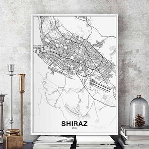 Op de afbeelding: Een zwart-witte kaartprint van Shiraz, Iran. De kaart toont de straten en bezienswaardigheden van de stad. De print is ingelijst in een wit frame en hangt aan een muur.
