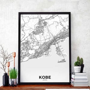 Puede incluir: Impresión de mapa en blanco y negro de Kobe, Japón en un marco negro. El mapa muestra las calles y la costa de la ciudad. El texto "KOBE JAPÓN" está impreso debajo del mapa.