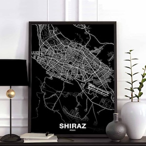 Op de afbeelding: Zwart-witte stadskaartprint van Shiraz, Iran. De kaart is ingelijst in een zwart frame en wordt tentoongesteld op een plank met een zwarte lamp en een witte vaas met een groene plant.