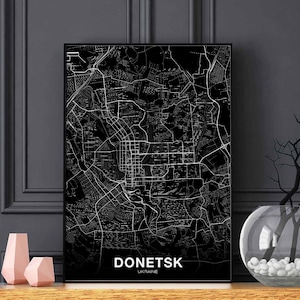 Puede incluir: Impresión de mapa de la ciudad de Donetsk, Ucrania, en blanco y negro. El mapa está enmarcado en un marco negro y se muestra en un estante con un jarrón y dos objetos decorativos de color rosa.