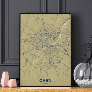 Puede incluir: Una impresión de mapa beige y azul de Caen, Francia, en un marco negro. El mapa muestra las calles y los puntos de referencia de la ciudad. El nombre de la ciudad está impreso en azul en la parte inferior del mapa.