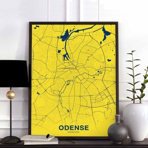 May include: A yellow and blue map print of Odense, Denmark, framed in a black frame. The print is hanging on a white wall with a black lamp and a plant in a white vase on a table below.