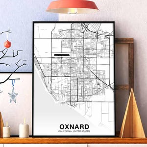 Może przedstawiać: Czarno-biały wydruk mapy Oxnard, Kalifornia, Stany Zjednoczone. Mapa przedstawia ulice i dzielnice miasta. Wydruk jest oprawiony w czarną ramę i wisi na ścianie.