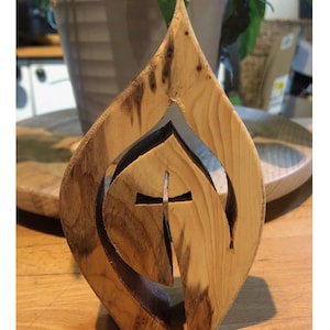 May include: A wooden sculpture of a flame with a cross inside. The sculpture is made of light brown wood with dark brown accents.