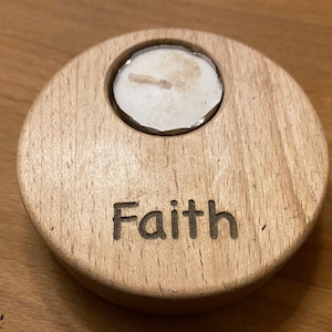 May include: A circular light-coloured wooden tea light candle holder with a smooth grain finish. The word Faith is engraved in a dark sans-serif font on the front surface. A single white tea light candle is inserted into the top circular cutout.