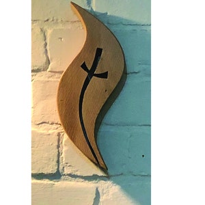 May include: A wooden flame-shaped wall decor with a black cross cut-out. The decor is mounted on a white brick wall.