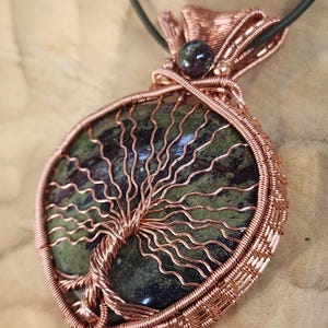 May include: A round pendant featuring a polished green and dark mottled stone wrapped in copper wire. The wire is intricately woven to form the shape of a tree with spreading branches across the stone surface, suspended from a dark cord.