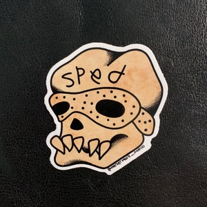 May include: A sticker of a skull wearing a mask with the word "Speed" written on the top. The sticker is on a black background. The sticker is a traditional tattoo style.