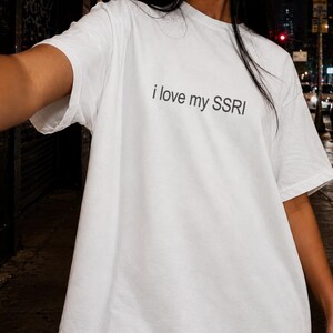 May include: A white short-sleeved t-shirt featuring the black text i love my SSRI printed in a simple sans-serif font across the chest. The garment has a crew neckline and a relaxed, loose-fitting silhouette with standard hemmed sleeves.