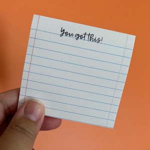 May include: A square off-white sticky note featuring horizontal blue lines and vertical red margin lines. The text "You got this!" is printed in black cursive script at the top of the note, which is held against a solid orange background.