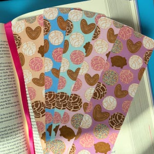 May include: A collection of colorful bookmarks with various patterns, including hearts, conchas, and pig-shaped designs. The bookmarks are made of paper and have a pink ribbon attached. They are placed on top of an open book.