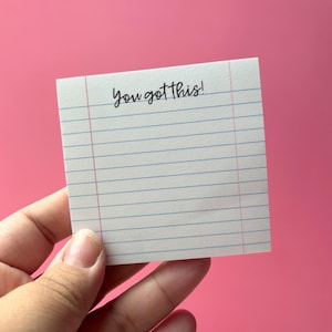 以下が含まれることがあります： 「You got this!」の文字が黒字で書かれた、白い罫線入りのメモ帳。メモ帳には青い横線と赤い縦の余白線があります。ピンク色の背景に配置されています。