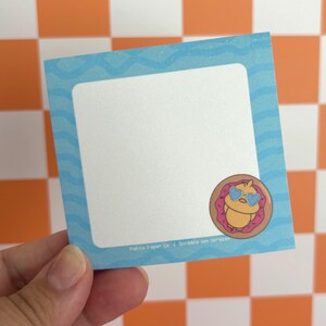 May include: A square sticky note with a light blue border featuring a wavy pattern and a white center. In the bottom right corner, there is a circular graphic of a yellow chick wearing heart-shaped glasses inside a pink donut. Text reads Patita Paper Co. | Scribble con corazon.