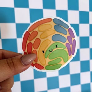 May include: A colorful sticker featuring a smiling, cartoon-style design. The sticker has a circular shape with a light tan background and is filled with rainbow-colored shapes. The sticker is held against a blue and white checkered background.