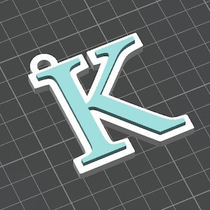 May include: A three-dimensional letter K pendant featuring a light blue face layered over a thicker white base. The letter has a small circular loop at the top left corner for attachment, presented against a dark gray grid background.