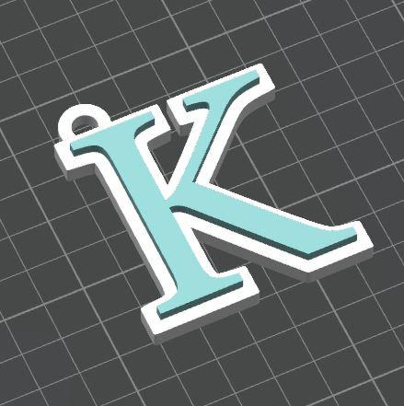 May include: A three-dimensional letter K pendant featuring a light blue face layered over a thicker white base. The letter has a small circular loop at the top left corner for attachment, presented against a dark gray grid background.