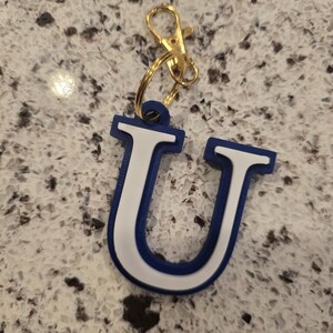 May include: A letter U keychain featuring a bold blue outer border and a white inner section. The letter is attached to a gold-toned metal split ring and a lobster claw clasp, providing a functional way to secure keys or attach to personal items.