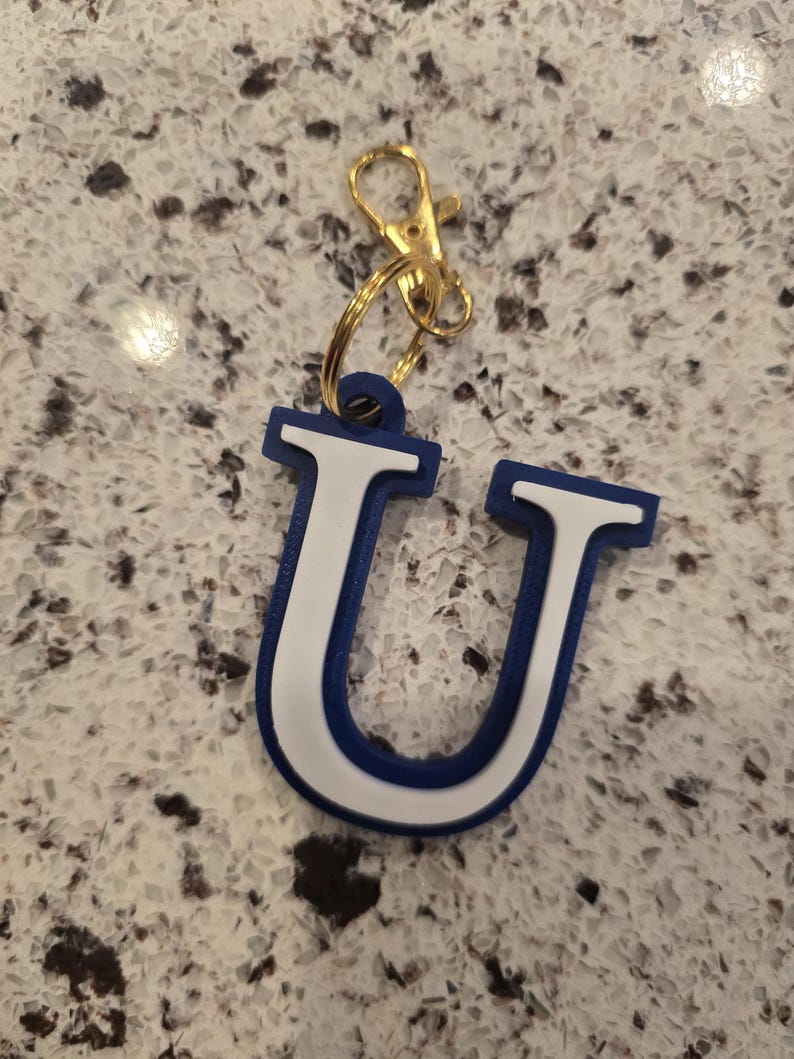 May include: A letter U keychain featuring a bold blue outer border and a white inner section. The letter is attached to a gold-toned metal split ring and a lobster claw clasp, providing a functional way to secure keys or attach to personal items.