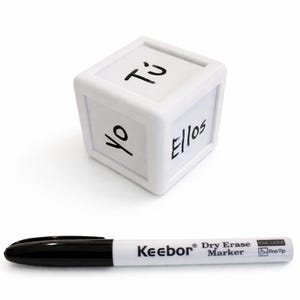 May include: A white cube with the words "Yo", "Tú", and "Ellos" written on its sides. A black and white Keebor dry erase marker is below the cube. The marker has a fine tip.