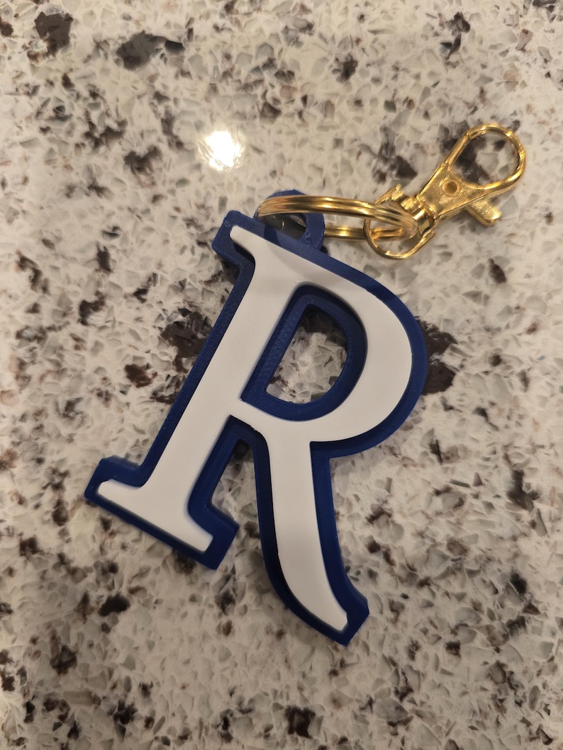 May include: A letter R keychain featuring a white letter with a thick blue border. The letter is attached to a gold-colored metal split ring and a matching gold-colored lobster claw clasp, providing a functional way to secure keys or accessories.
