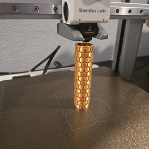 Puede incluir: Una impresora 3D está creando un objeto cilíndrico dorado con un patrón de panal. El cabezal de la impresora, blanco y gris, muestra el logotipo "Bambu Lab". El objeto se imprime sobre una superficie oscura.