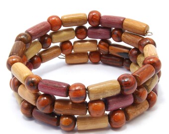 Puede incluir: Un brazalete de madera multicolor con cuentas redondas y cilíndricas. El brazalete está hecho de diferentes tonos de madera marrón, roja y morada.