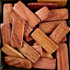 May include: A pile of rectangular wooden pieces with a reddish-brown color and a wood grain pattern. The pieces are arranged in a random order and some are overlapping.