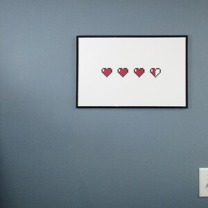 May include: A framed print with a white background and five red pixelated hearts. The hearts are arranged in a row, with the last heart partially filled in.