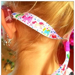 Puede incluir: Una niña pequeña con una correa para audífonos con temática de unicornio blanca y rosa. La correa está unida a un audífono detrás de su oreja. También lleva un pendiente con un dije de unicornio.