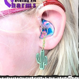 May include: A close-up of a hearing aid with a green cactus charm attached to it. The hearing aid is in a person's ear. The charm is hanging from a gold chain with green beads.