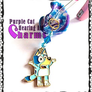 May include: A hearing aid charm featuring a cartoon dog in shades of blue, white, and gold. The charm is attached to a blue and purple marbled hearing aid mold with a clear tube. The text "Purple Cat Hearing Aid Charms" is visible.