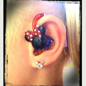 May include: A close-up of a child's ear with a hearing aid decorated with a red and white Minnie Mouse design. The hearing aid is attached to the ear with a clear, plastic mold. The child is also wearing a pair of gold Mickey Mouse earrings.