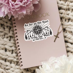 May include: A pink notebook with the text "NOTEBOOK" and a sticker that reads "As far as the East is from the West" sits on a cream-colored knitted blanket. A gold necklace with a cross and two peonies, one pink and one white, are also in the image.