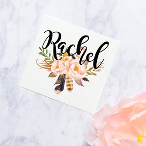 May include: A white card with the name "Rachel" written in black cursive script. The name is surrounded by a floral design with pink and orange flowers, green leaves, and brown feathers.